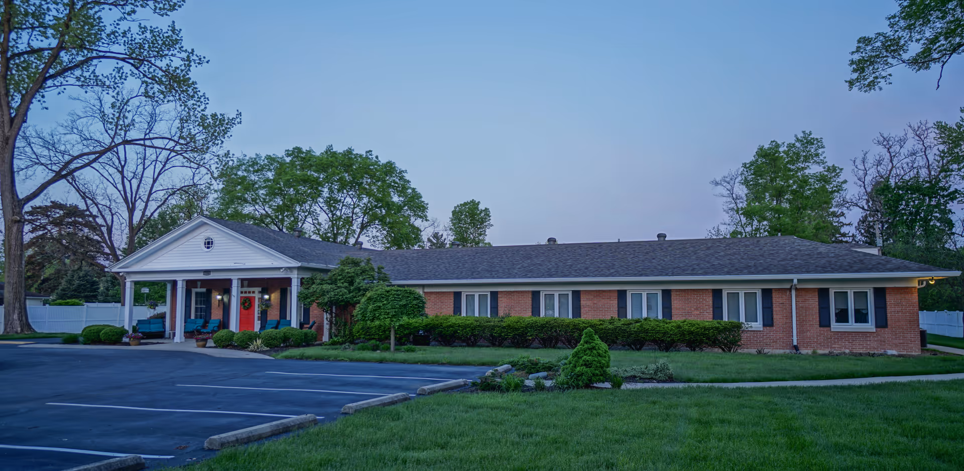 Single-story brick senior living building with a covered columned entrance, red front door, parking lot, and landscaped lawn.