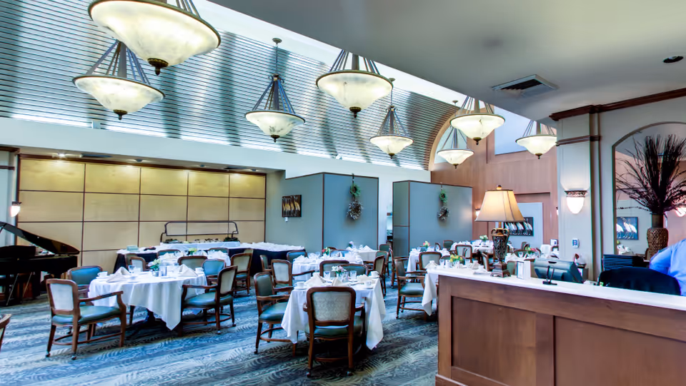 A spacious dining room with multiple round tables covered in white tablecloths, each set with plates, glasses, and silverware. The room features elegant hanging light fixtures, a grand piano in the corner, and a reception desk with a lamp. The decor includes wall art and plants, creating a welcoming atmosphere.