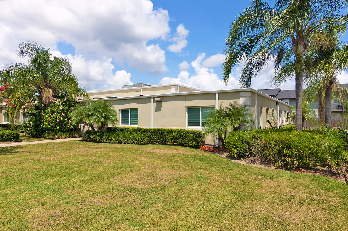 Low single-story beige building with palm trees, hedges and a grassy lawn under a partly cloudy blue sky.