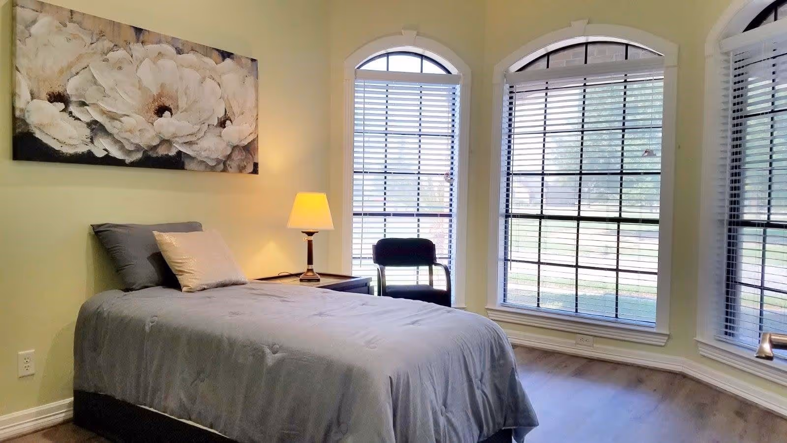 Sunlit bedroom with a made bed, bedside table and lamp, a chair, and three tall arched windows with blinds.