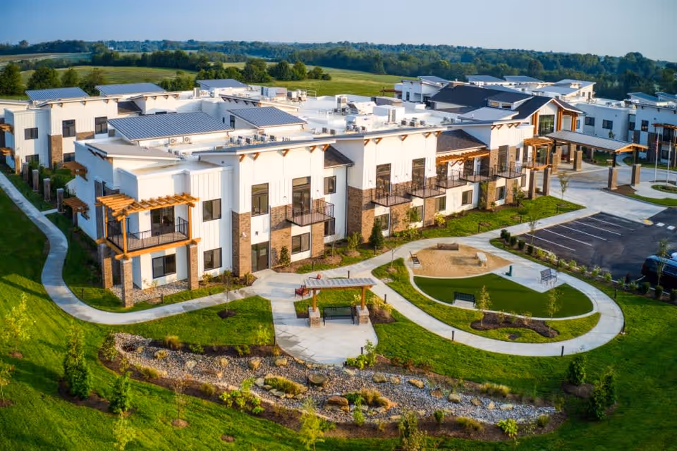 Aerial view of Preston Greens Senior Living facility showing a modern two-story building with balconies, surrounded by well-maintained green lawns, walking paths, benches, and a small landscaped garden area. The parking lot is visible on the right side, and the background features open fields and trees under a clear sky.