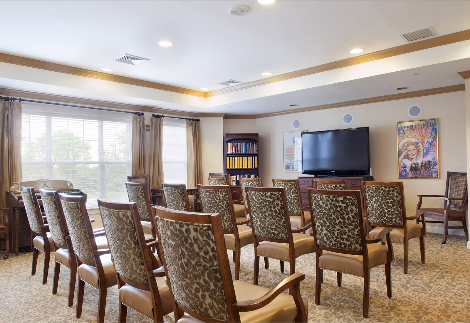 Sunlit activity room with rows of patterned chairs facing a large flat-screen TV and bookshelves.