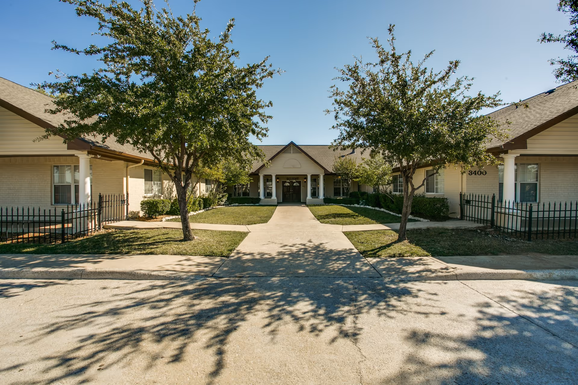 Front exterior view of Avalon Memory Care facility with a paved walkway leading to the entrance, flanked by two trees and green lawns on either side under a clear blue sky.