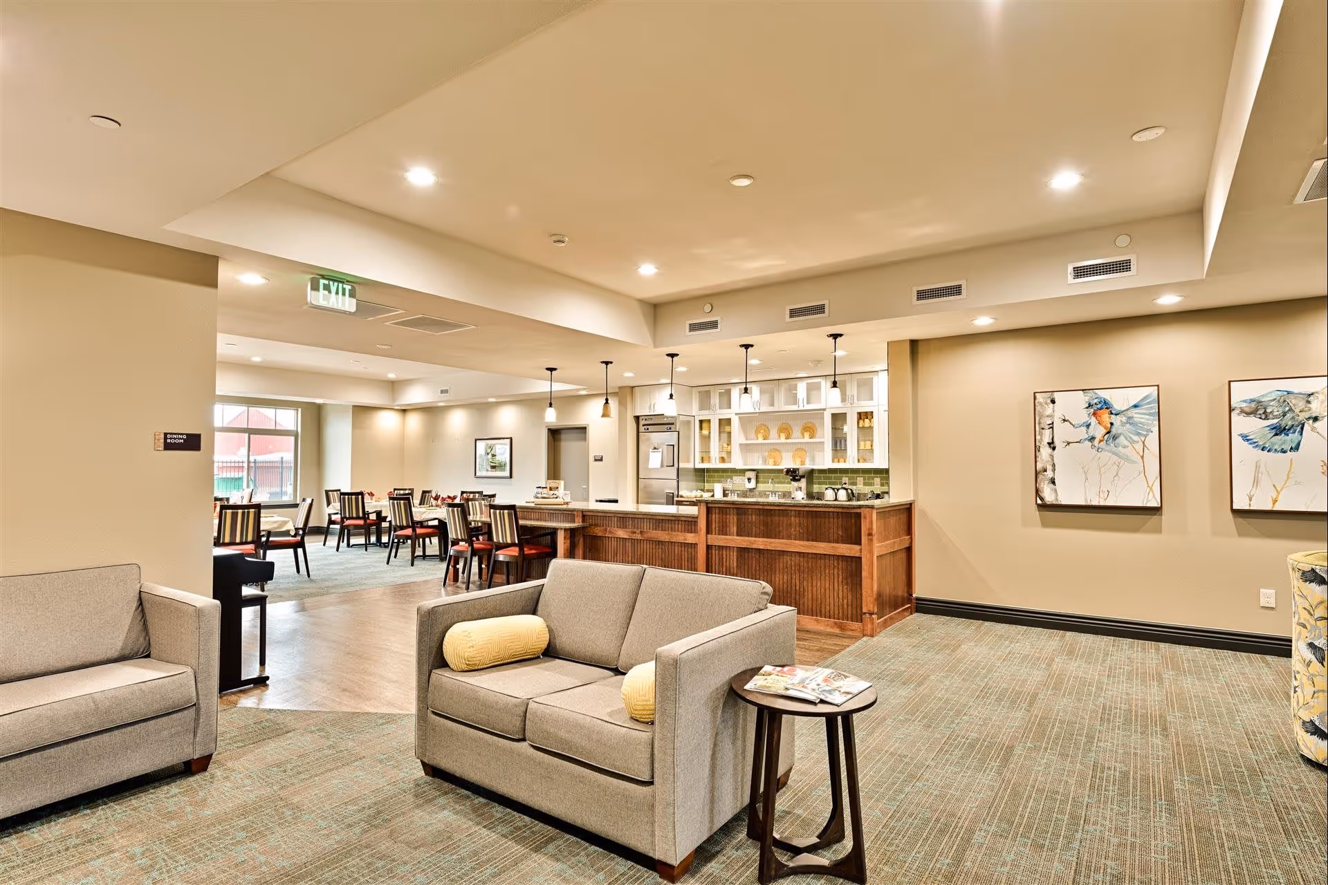 Interior view of a senior living facility common area with beige walls and carpeted floor. The foreground features two gray sofas with yellow cylindrical pillows and a small round side table with magazines. In the background, there is a dining area with multiple tables and chairs, and a kitchen area with a wooden counter, pendant lights, and white cabinets. Two framed bird paintings hang on the wall to the right.