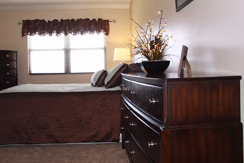 Sunlit bedroom with a bed, dark wood dresser topped with a lamp and decorative arrangement, and a window with a valance.