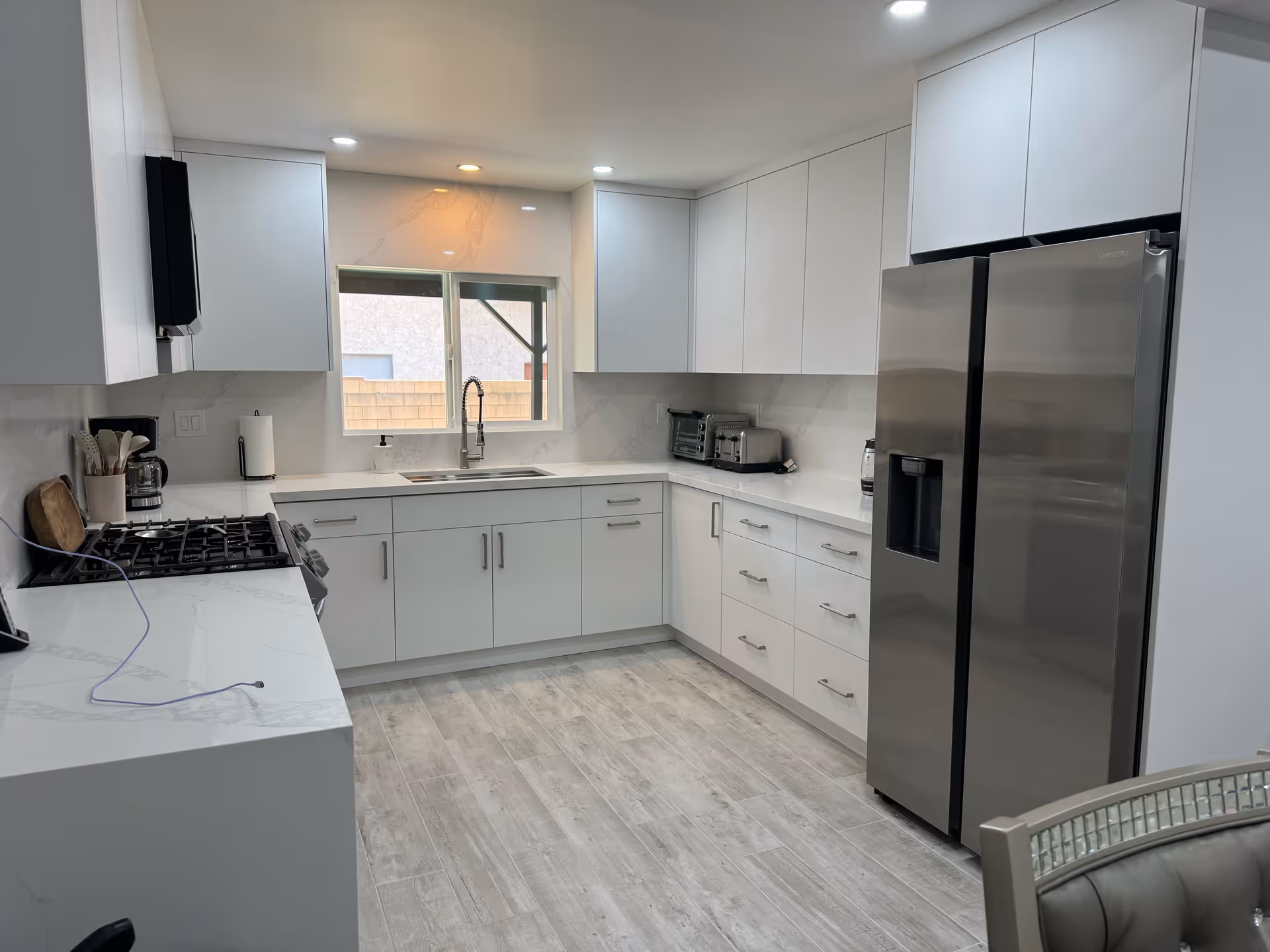 Modern white U-shaped kitchen with stainless steel refrigerator, gas range, sink under a window, and light wood-look flooring.