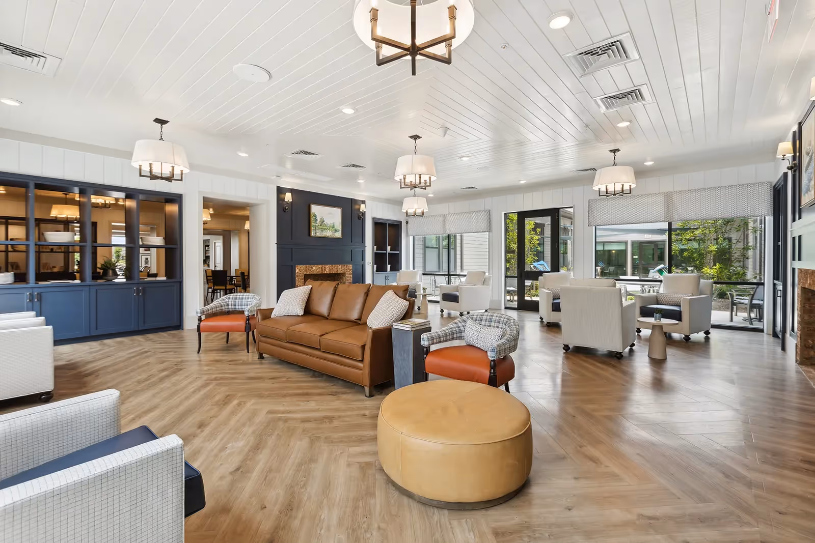 A spacious and well-lit living room area in a senior living facility featuring a brown leather sofa, several armchairs in light and plaid patterns, a round tan ottoman, and multiple modern chandeliers. Large windows and glass doors provide natural light and views of an outdoor patio area with greenery. The room has wood flooring, white paneled walls, and built-in blue cabinetry with decorative items.