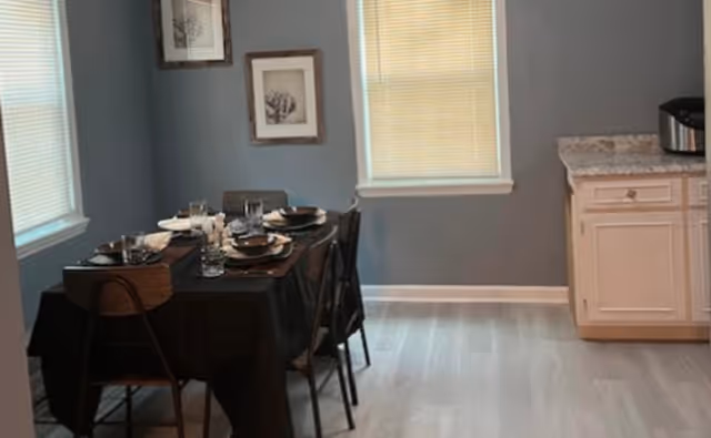 A dining area with a rectangular table covered with a black tablecloth set for six people with plates, glasses, and cutlery. The room has gray walls, two windows with blinds, two framed pictures on the wall, and a white cabinet with a granite countertop on the right side.