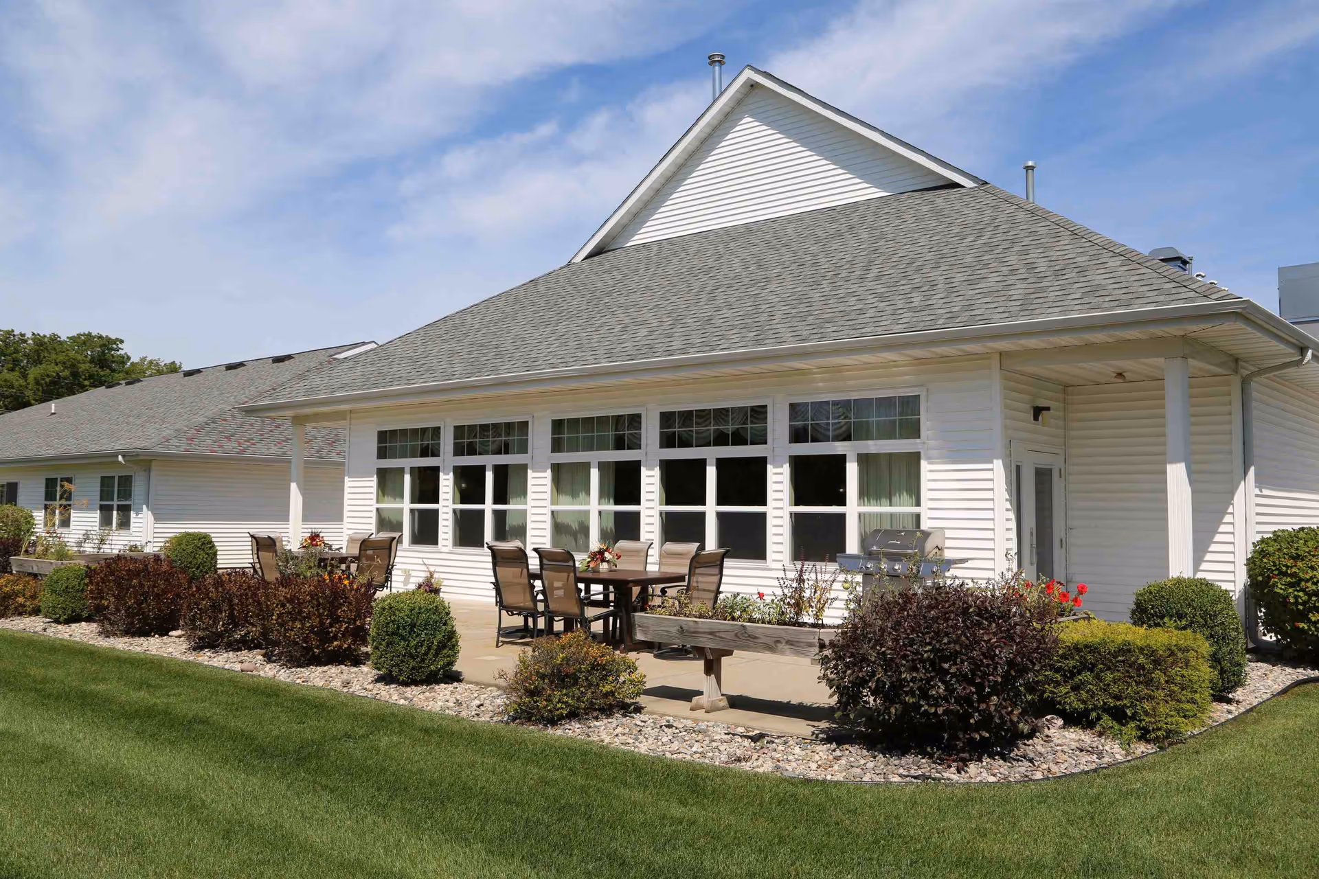 White single-story senior living building exterior with a patio, outdoor table and chairs, and landscaped lawn.