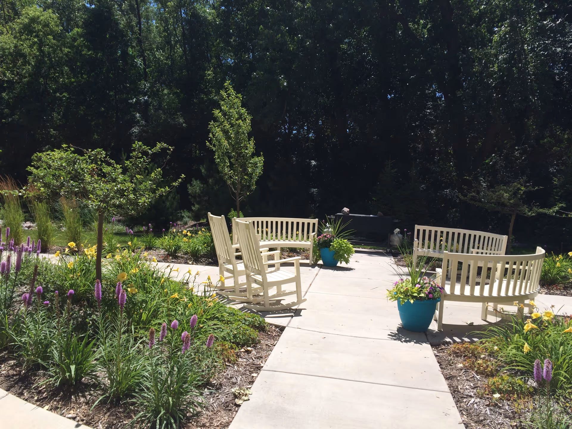 Outdoor garden area with a concrete pathway surrounded by flower beds containing purple and yellow flowers. There are white wooden benches and rocking chairs arranged around the pathway, with trees and dense greenery in the background.