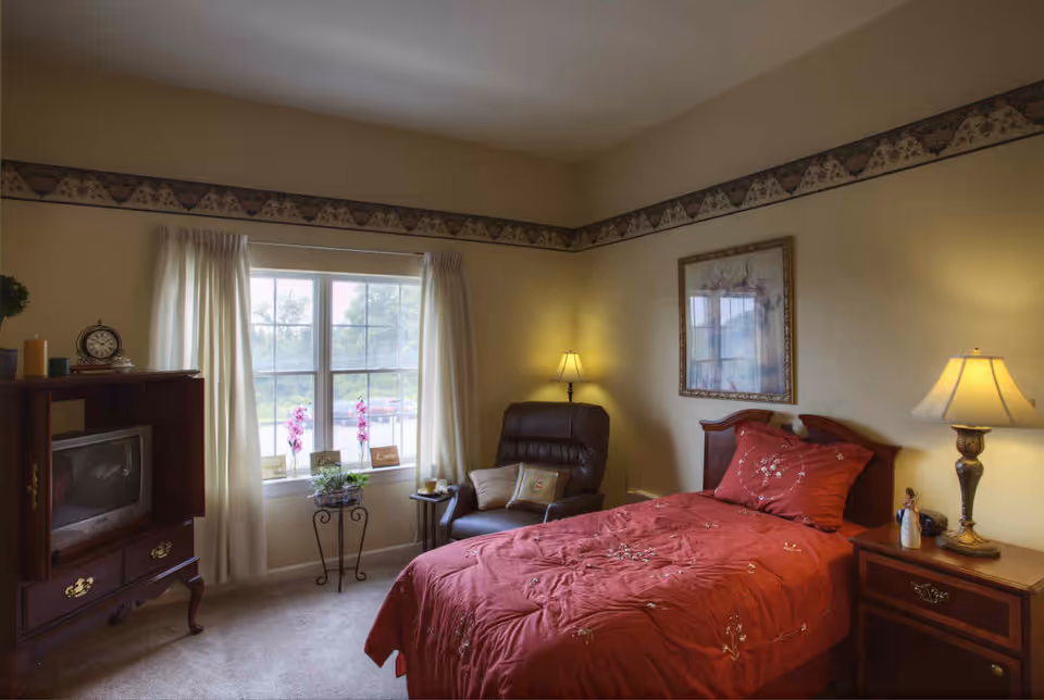 A cozy bedroom with a single bed covered in a red floral comforter, a wooden nightstand with a lamp, a comfortable armchair with pillows, a window with white curtains letting in natural light, and a wooden cabinet with a TV on the left side.