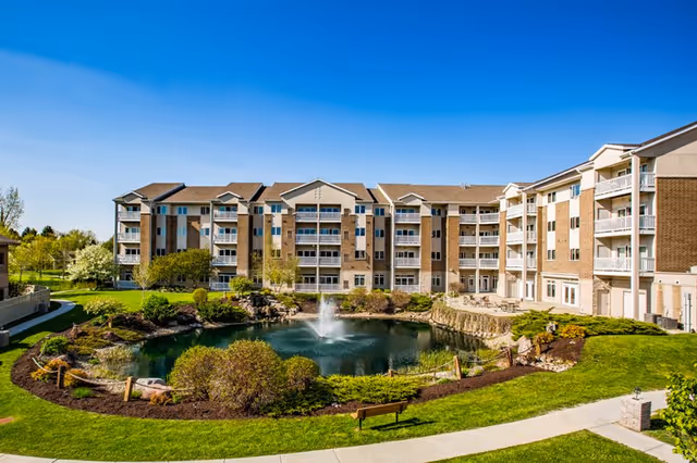 Exterior view of a multi-story senior living facility named Coventry Village with balconies, surrounded by well-maintained landscaping including a pond with a water fountain, green grass, bushes, and trees under a clear blue sky.