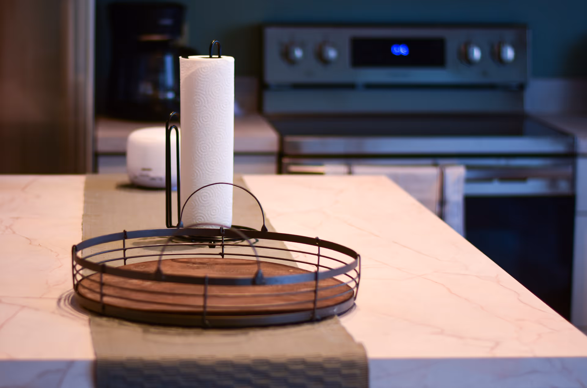 Close-up view of a kitchen countertop with a paper towel holder and a round metal tray with a wooden base. In the background, there is a stove with a digital clock and a coffee maker on the counter.