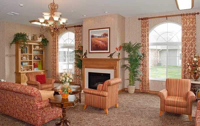 A cozy living room area with patterned carpet and wallpaper. The room features a wooden fireplace with a framed landscape painting above it, two large arched windows with floral curtains, and several upholstered chairs and a sofa in red and orange tones. There is a wooden bookshelf with decorative items and books, a small round wooden coffee table with a flower arrangement, and potted plants adding greenery to the space.