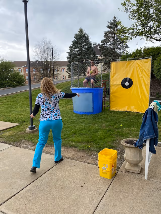 A woman in blue pants and a floral top is throwing a ball at a man sitting in a dunk tank outdoors on a grassy area. The man is shirtless and wearing shorts, sitting inside a blue tank with a yellow target panel beside it. There are trees, a building, and a sidewalk visible in the background.