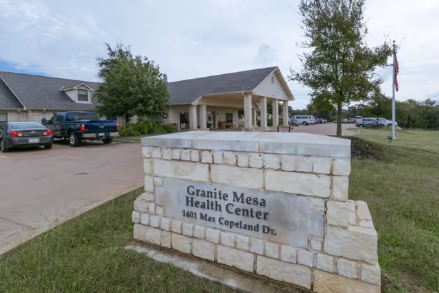 Exterior view of Granite Mesa Health Center showing a stone sign with the facility's name and address, a driveway with parked cars, and the entrance to the building with a covered porch and columns.