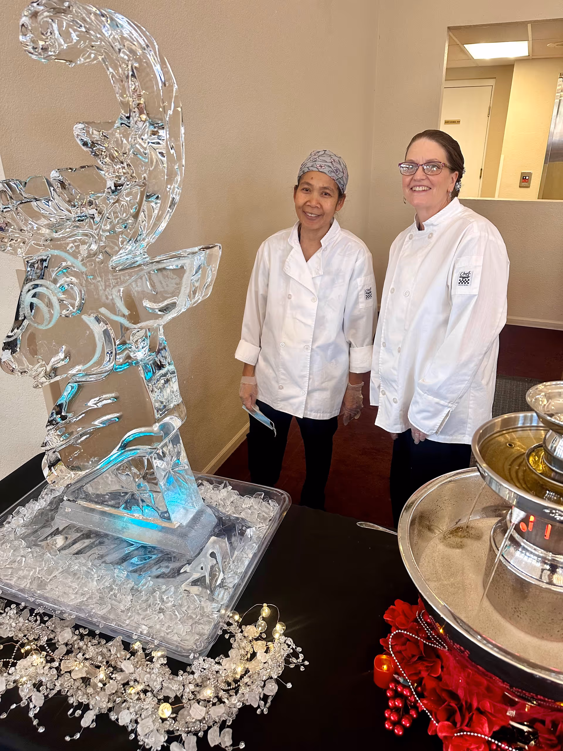 Two women wearing white chef coats stand indoors next to an ice sculpture of a reindeer and a chocolate fountain decorated with red flowers and beads.