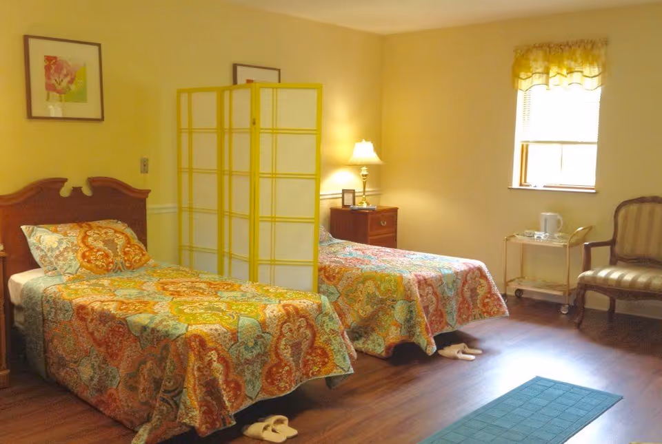 Two twin beds with colorful patterned bedspreads in a well-lit bedroom with a folding room divider, nightstand, lamp and chair.