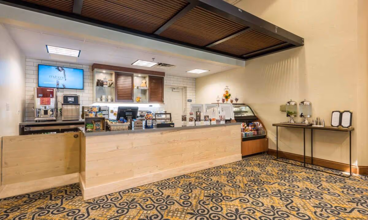 Interior view of a small café or snack bar area with a wooden counter, a coffee machine, a display case with snacks and drinks, a patterned carpet, and a small table with water dispensers and menus against a beige wall.