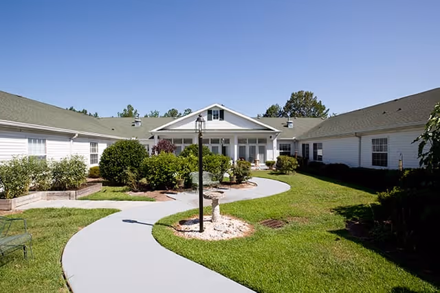 A sunny outdoor courtyard area with a curved concrete walkway, green grass, bushes, and a lamppost in the center. The courtyard is surrounded by a single-story white building with a green roof and multiple windows.