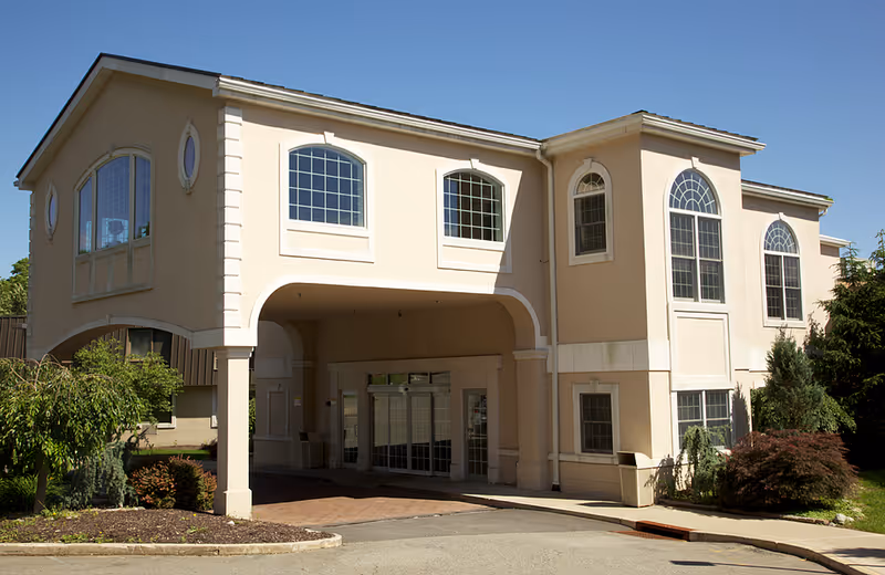 Exterior view of a two-story beige building with large arched windows and an entrance covered by a portico. There are shrubs and small trees around the building under a clear blue sky.