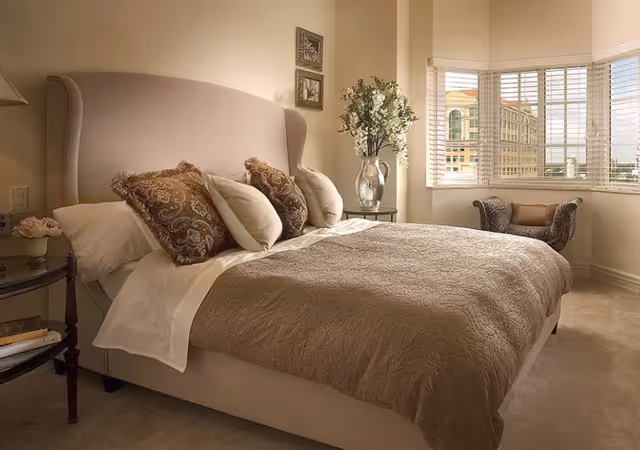 A cozy bedroom with a large beige upholstered bed adorned with multiple decorative pillows and a textured bedspread. Next to the bed is a small round side table with a lamp, a small flower arrangement, and some books. A large window with white blinds allows natural light to fill the room, and a patterned armchair with a cushion is placed near the window. A vase with white flowers sits on a small table in the corner.