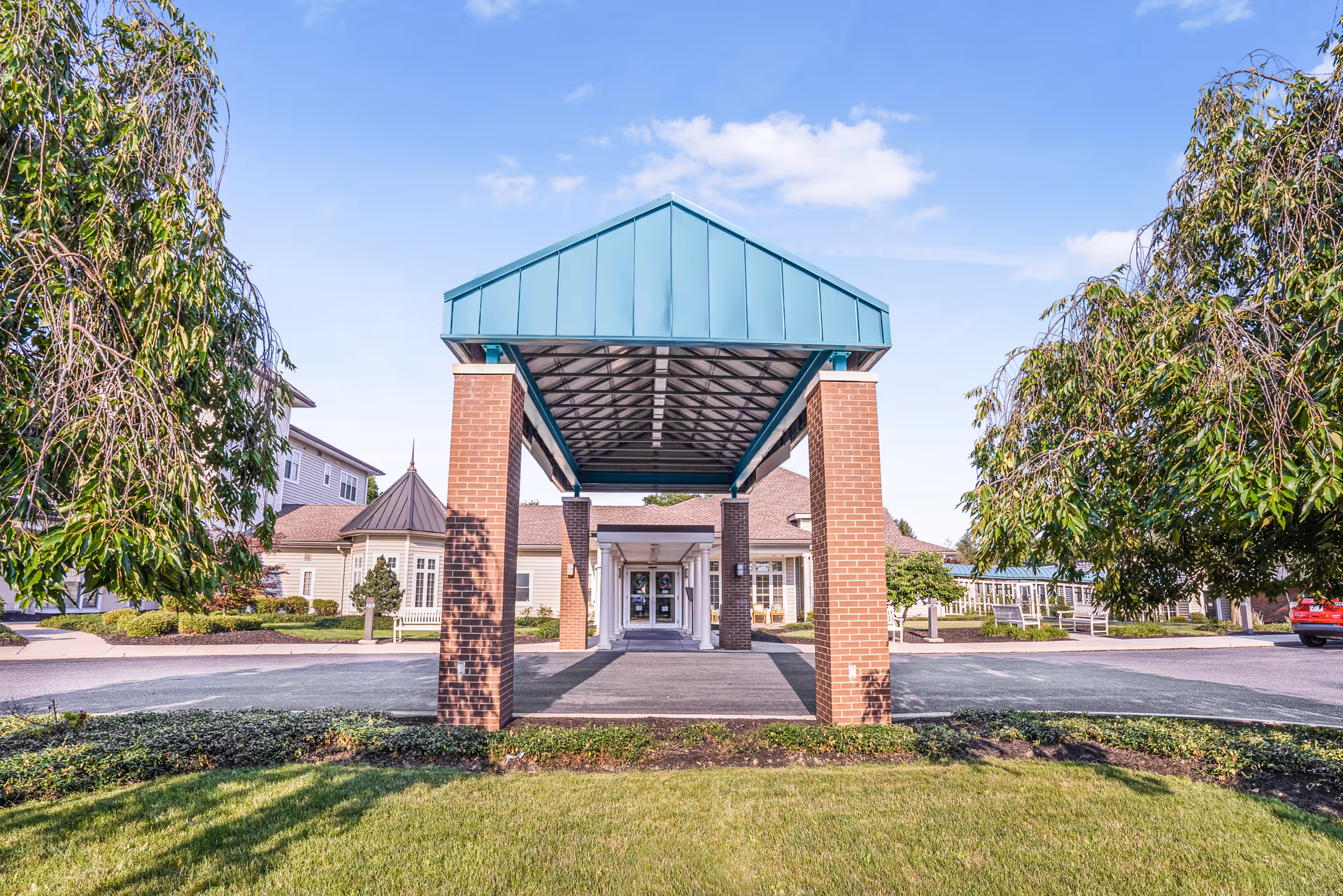 Front entrance of a senior living facility featuring a covered porte-cochere with brick columns and landscaped grounds.