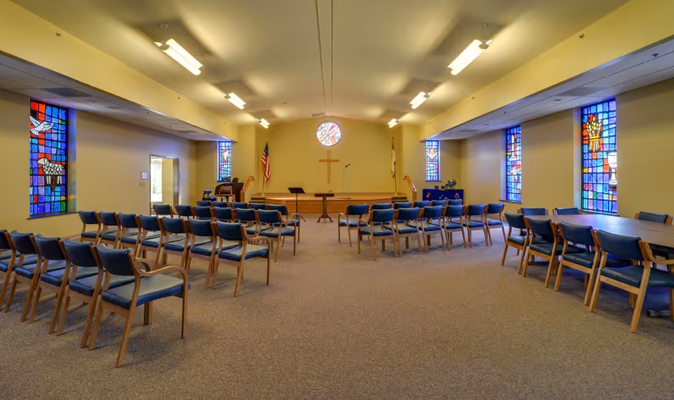 Interior view of a chapel or worship room with rows of blue cushioned wooden chairs arranged facing a small stage with a wooden cross on the wall, an American flag, a Christian flag, and colorful stained glass windows on both sides.