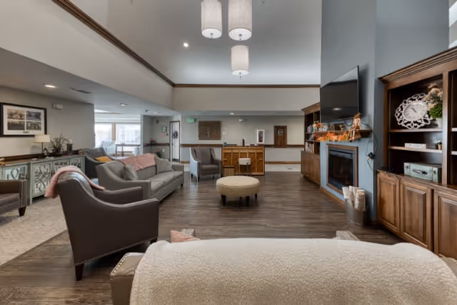 A spacious and well-lit living room area in a senior living facility featuring comfortable seating including sofas and armchairs, a round ottoman in the center, a wall-mounted flat-screen TV above a fireplace, wooden built-in shelves with decorative items, and a wooden floor. The room has a high ceiling with recessed lighting and a soft neutral color palette.