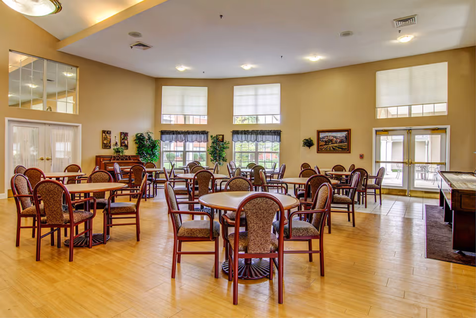 A spacious dining room with multiple round and rectangular tables surrounded by cushioned chairs. The room has large windows with white blinds and valances, beige walls, and wooden flooring. There are decorative plants and framed artwork on the walls, and double glass doors leading outside.