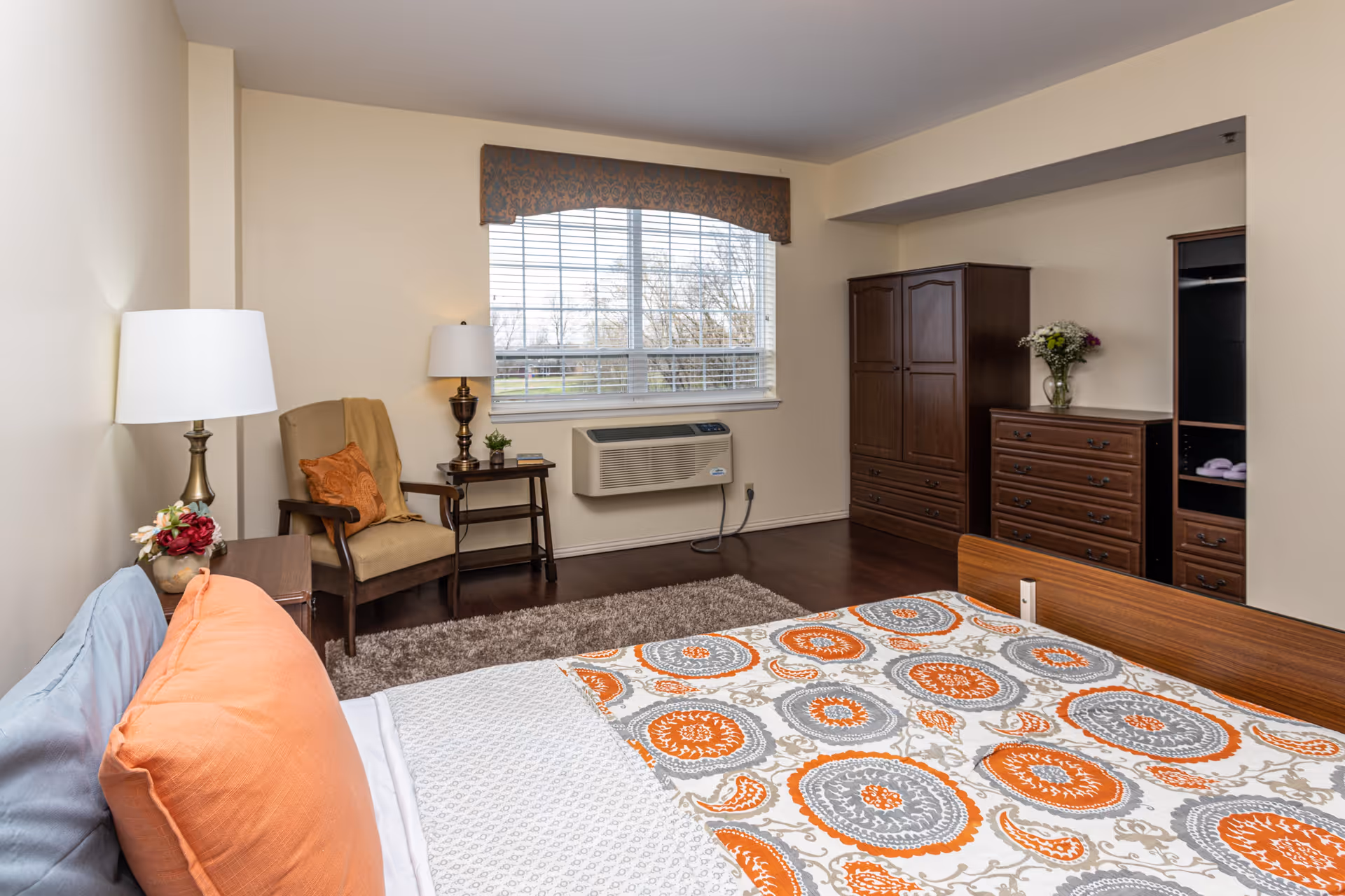 A cozy bedroom with a bed featuring a colorful orange, gray, and white patterned bedspread and orange pillows. There is a beige armchair with an orange cushion and a beige throw blanket next to a small wooden side table with a lamp and a small plant. A large window with blinds and a decorative valance lets in natural light. Against the far wall are a wooden wardrobe and a chest of drawers with a vase of flowers on top. The floor is dark wood with a light brown rug near the bed.