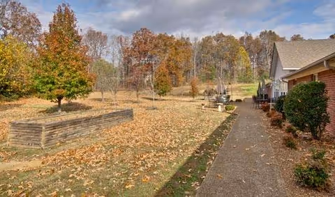 Outdoor area of a senior living facility with a paved walkway alongside a building. The ground is covered with fallen autumn leaves, and there are trees with fall foliage in the background. Raised garden beds and some outdoor furniture are visible.