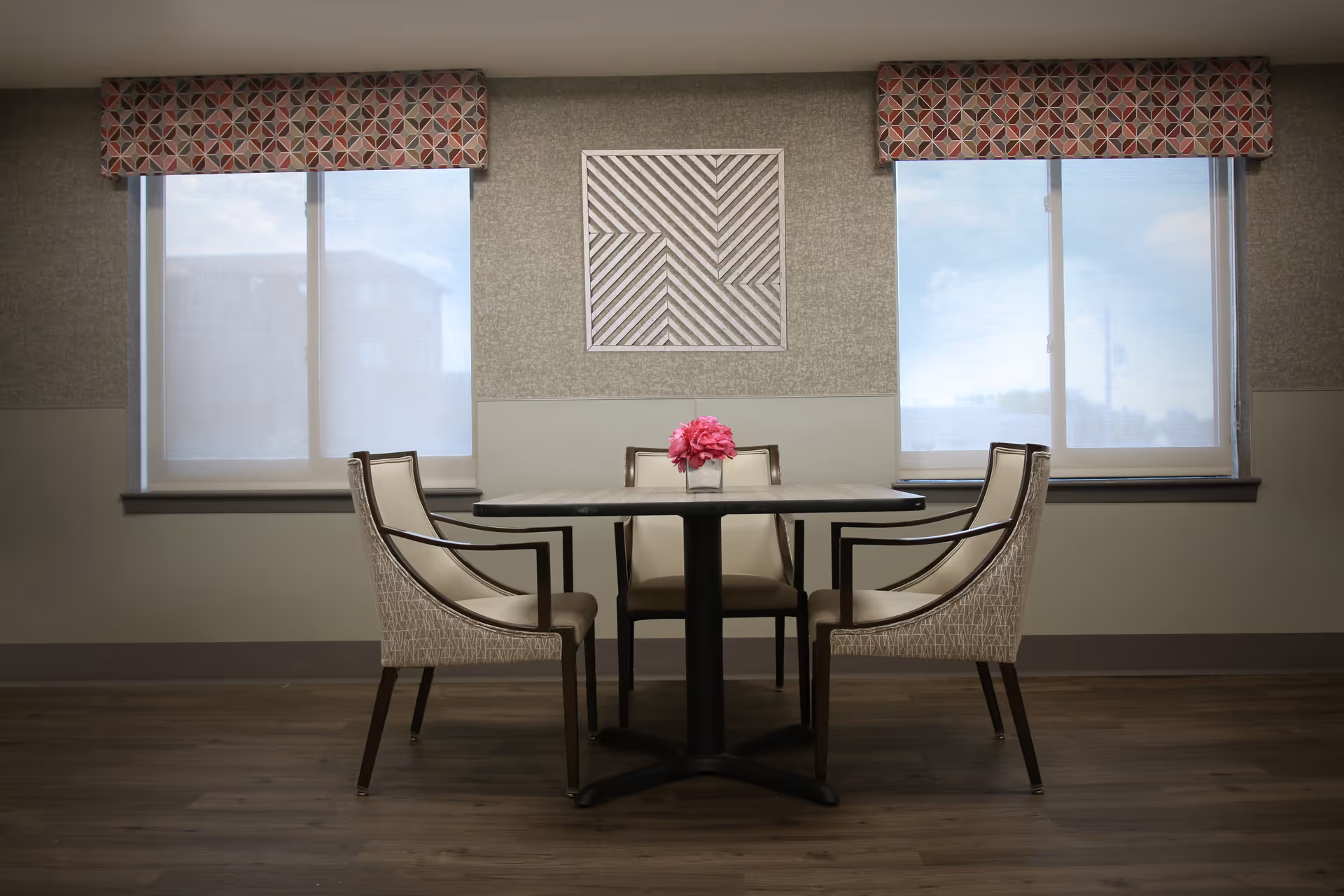A small dining area with a square table and four cushioned chairs arranged around it. The table has a small vase with pink flowers in the center. Behind the table are two large windows with patterned valances and light filtering shades. A geometric wall art piece is centered between the windows. The floor is wood and the walls are painted in neutral tones.
