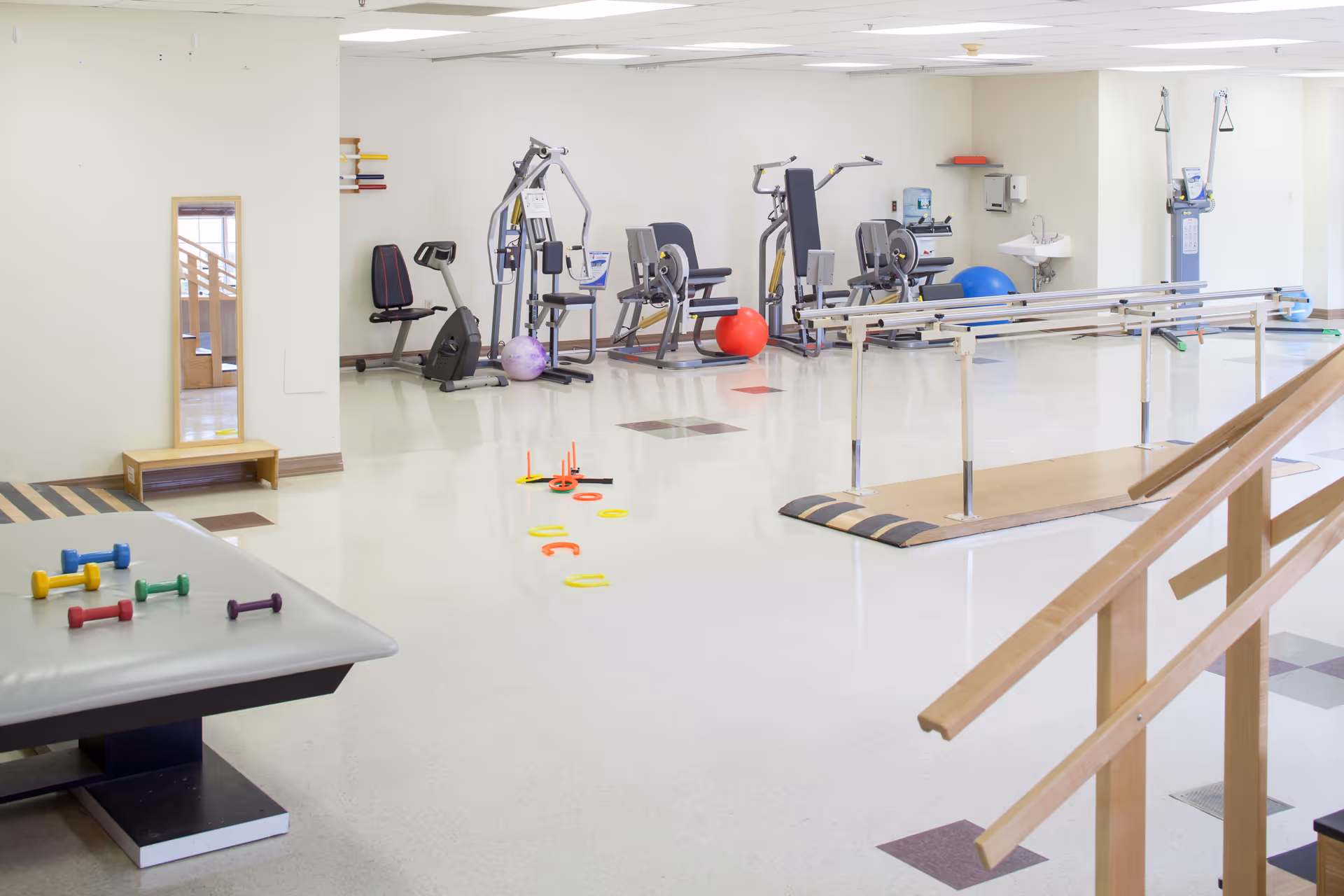 A bright physical therapy or rehabilitation room with various exercise equipment including stationary bikes, weight machines, colorful dumbbells on a padded table, balance balls, and parallel bars for walking exercises. The room has a clean, polished floor and a large mirror mounted on the wall.
