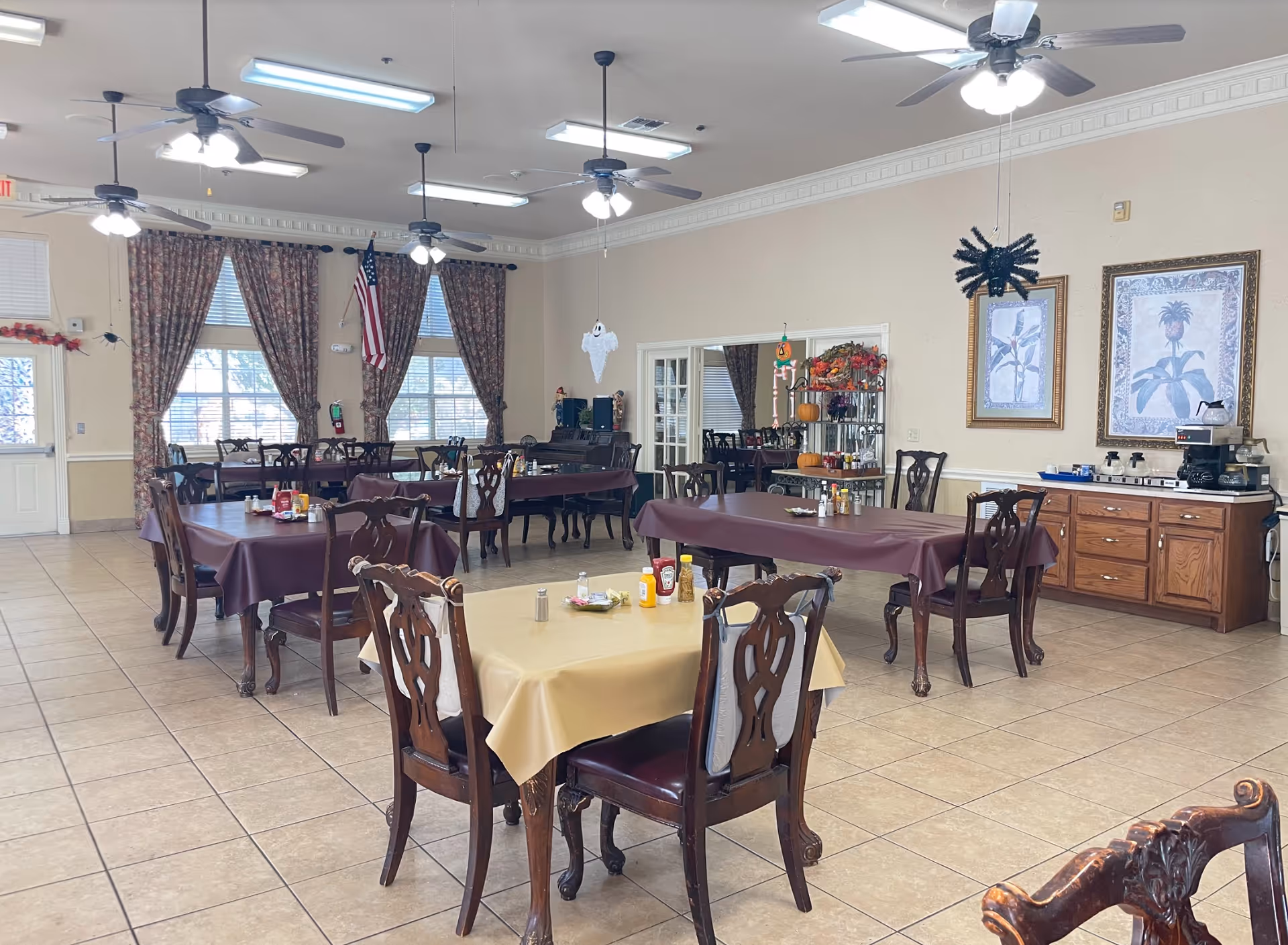 A spacious dining room with several wooden tables and chairs. Some tables are covered with maroon tablecloths and one table has a beige tablecloth. The room has tiled floors, ceiling fans with lights, and large windows with floral curtains. There are Halloween decorations hanging from the ceiling, including a ghost and a spider. On one side of the room, there is a wooden cabinet with a coffee maker and other items, and framed botanical artwork hangs on the wall.