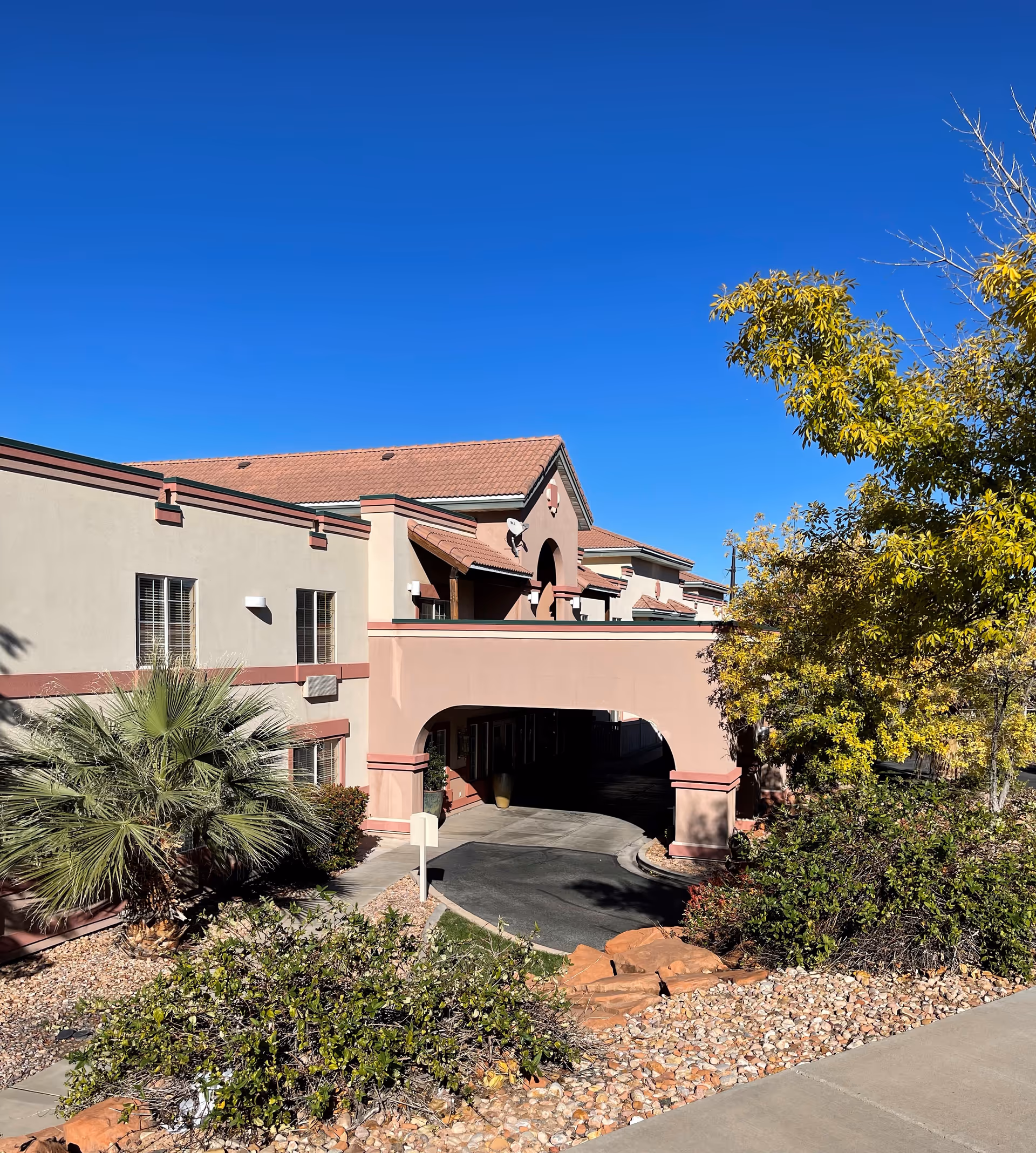 Exterior view of a senior living facility building with beige and pink walls, a covered driveway entrance, surrounded by desert landscaping including palm trees, bushes, and rocks under a clear blue sky.