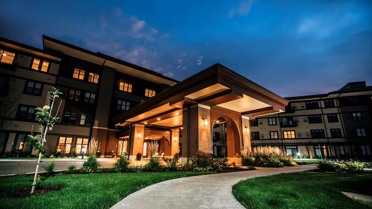 Exterior view of a senior living facility named Grand Living At Bridgewater at dusk, showing a well-lit entrance with a covered driveway, surrounded by landscaped greenery and a curved sidewalk.