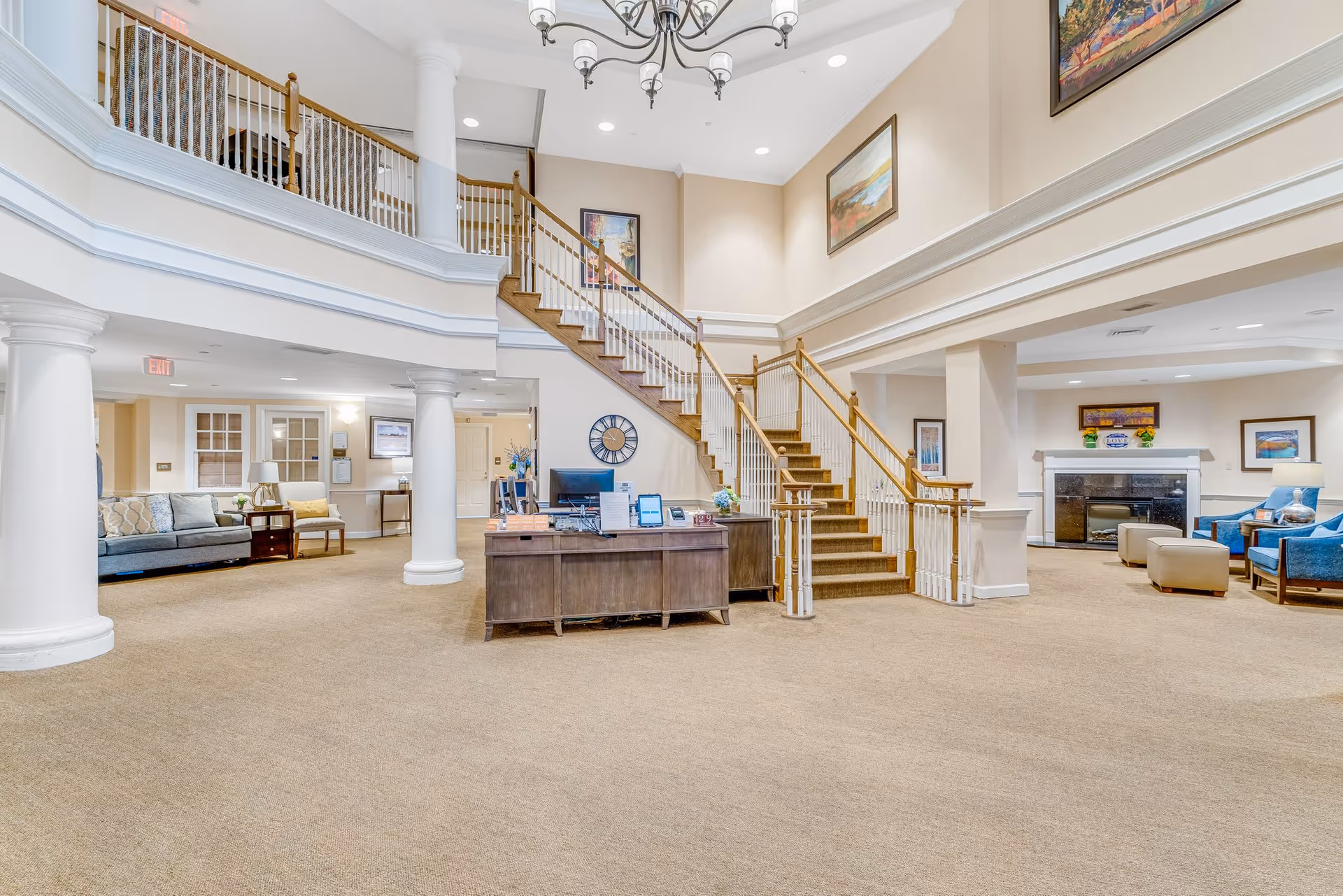 Spacious senior living facility lobby with a central wooden reception desk, a staircase with wooden handrails leading to an upper floor, seating areas with sofas and chairs, large white columns, beige carpet, and framed artwork on the walls.