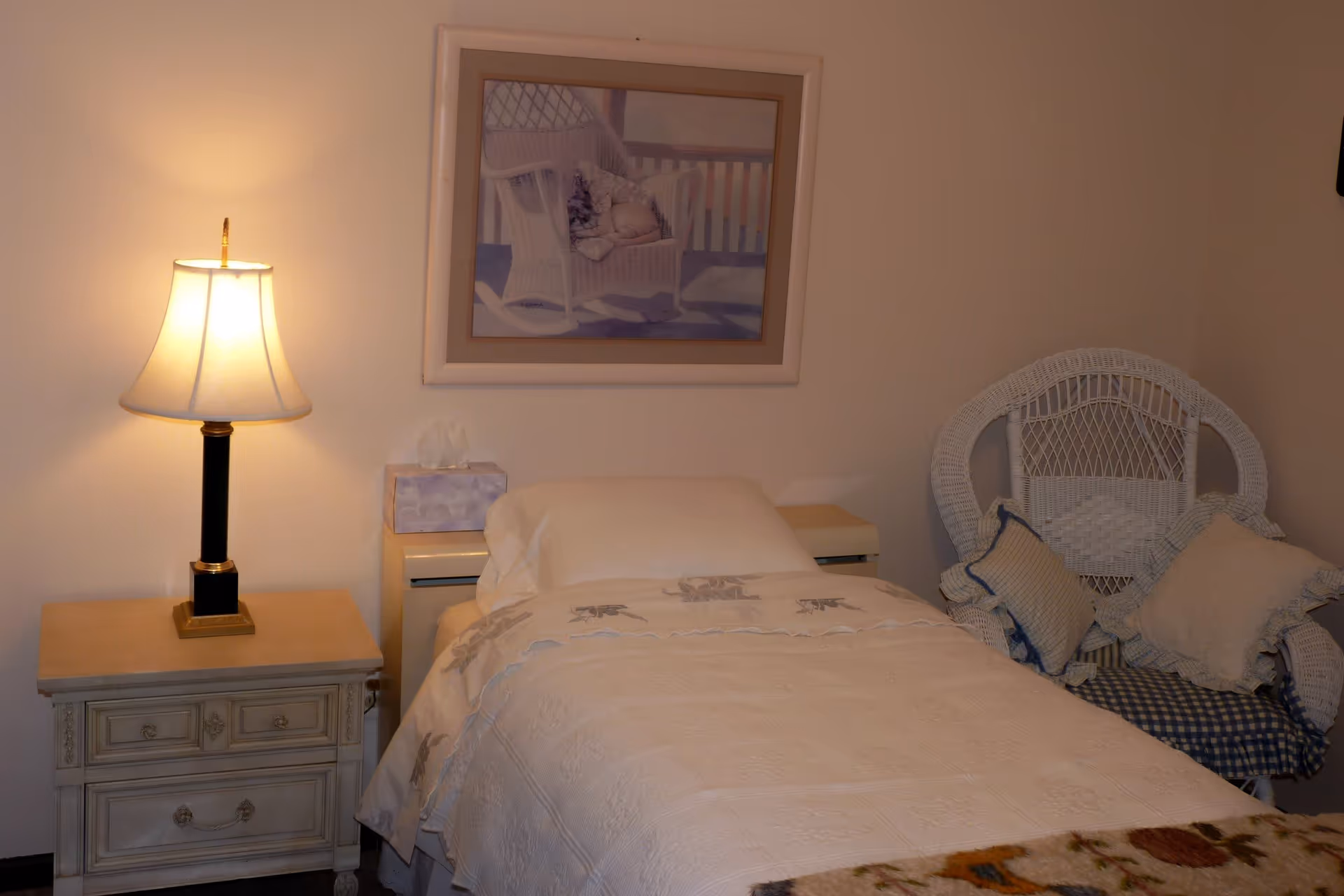 A cozy bedroom with a single bed covered in a white quilt, a bedside table with a lamp and a tissue box, a white wicker chair with cushions, and a framed picture of a rocking chair on the wall.