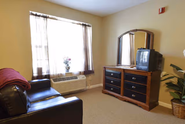 A cozy room with a black leather couch on the left, a window with sheer curtains letting in natural light, an air conditioning unit below the window, and a wooden dresser with a mirror and an old-style CRT television on top. There is a potted plant in a wicker basket on the right side of the dresser.