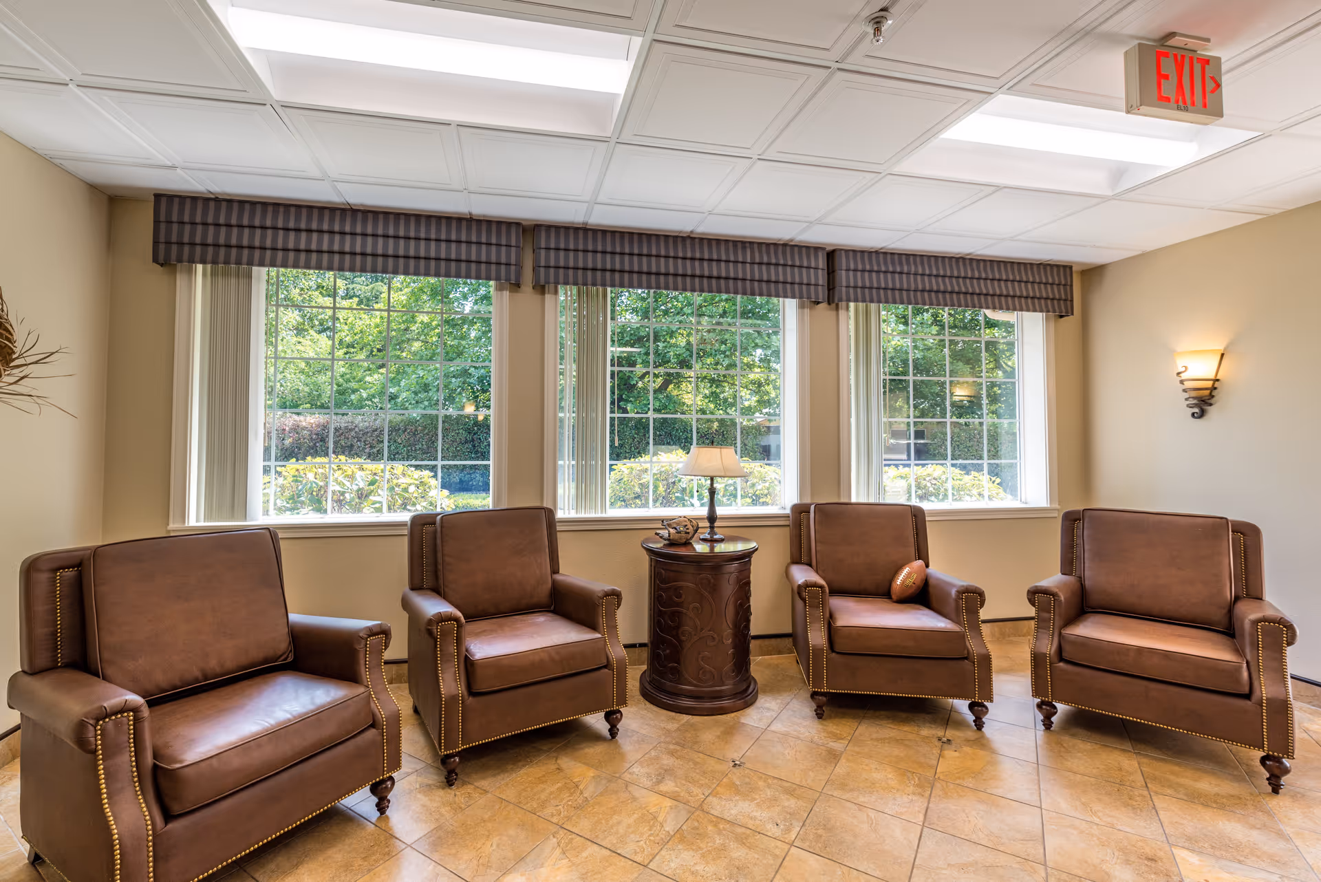 A bright sitting area with four brown leather armchairs arranged around a small round wooden table with a lamp on it. Large windows with striped valances let in natural light and show greenery outside. The floor is tiled, and there is a wall sconce and an exit sign visible.