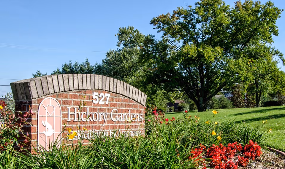 A brick sign for Hickory Gardens with the address number 527, surrounded by green grass, colorful flowers, and trees under a clear blue sky.
