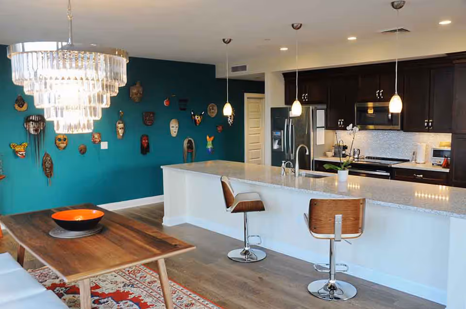 Modern kitchen and dining area with a wooden dining table, a large chandelier above it, two wooden bar stools at a white kitchen island with a sink, dark wood cabinets, stainless steel refrigerator, and a teal accent wall decorated with various colorful masks.