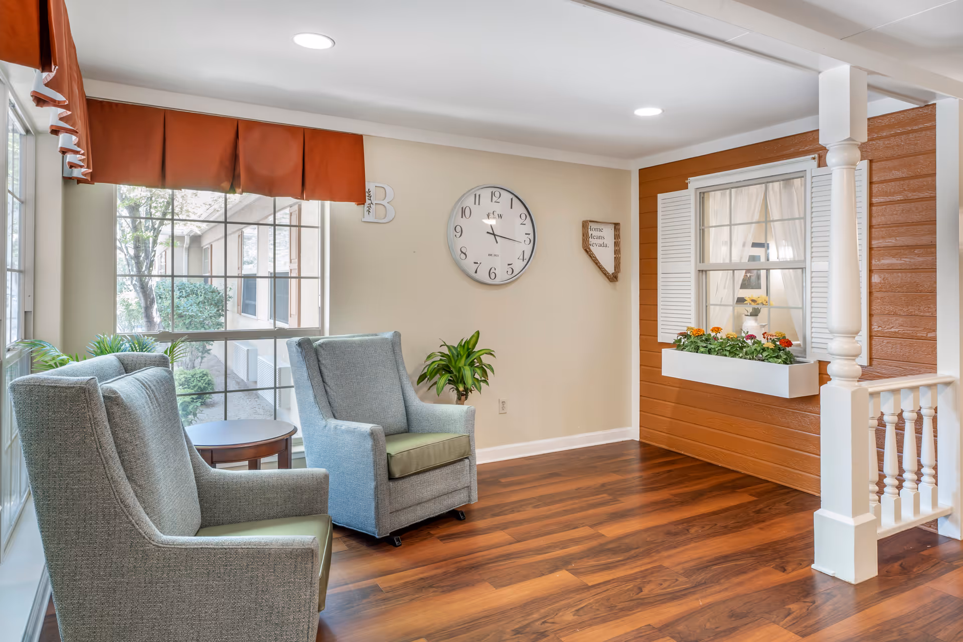A cozy sitting area with two gray armchairs with green cushions facing a large window with an orange valance. There is a small round wooden side table between the chairs and a green potted plant against a beige wall. A large round clock and a decorative sign shaped like Nevada with the words 'Home Means Nevada' hang on the wall. To the right, there is a section of wall designed to look like the exterior of a house with orange siding, white window shutters, a window box with flowers, and a white column and railing.