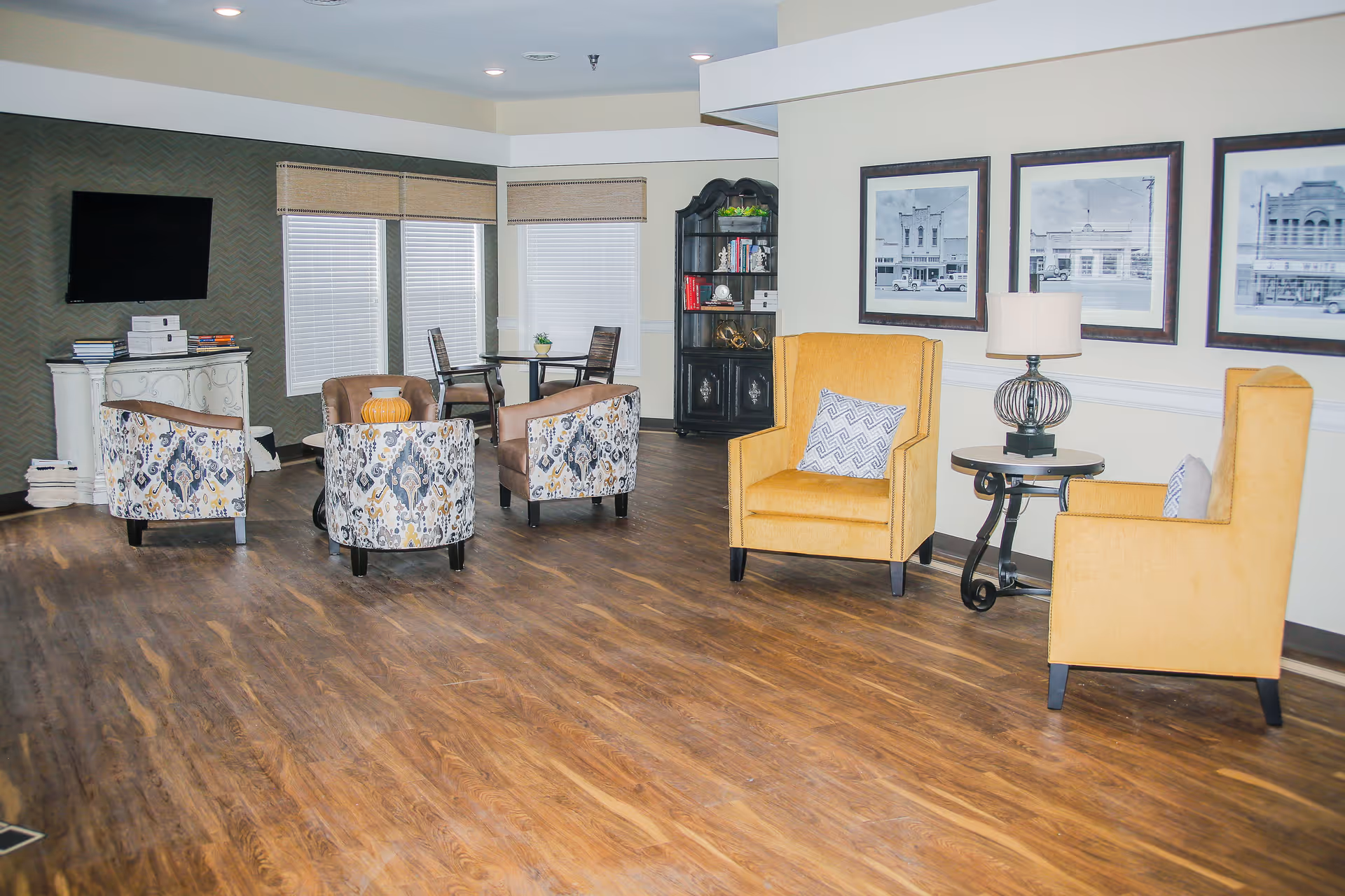 Spacious senior living common room with wooden floors, seating groups of patterned and yellow armchairs, a TV, bookshelf, and framed artwork on the wall.