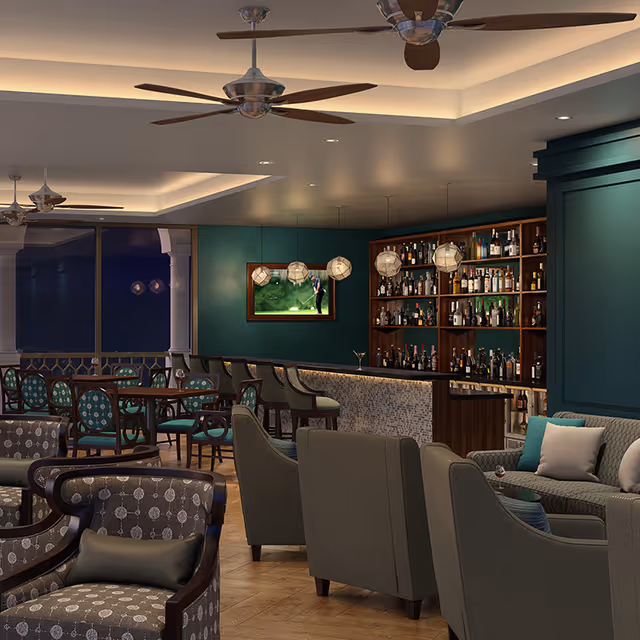 A stylish lounge area with comfortable armchairs and sofas arranged around small tables. In the background, there is a bar with shelves stocked with various bottles and hanging pendant lights. The walls are painted green, and a flat-screen TV is mounted on the wall showing a golf game. Ceiling fans and recessed lighting provide illumination.