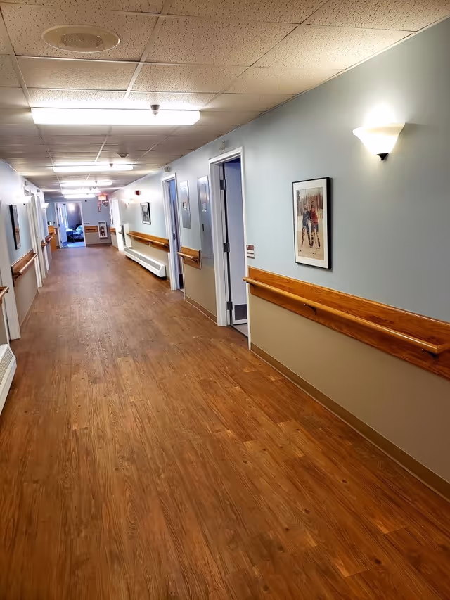 A long hallway in a senior living facility with wooden handrails on both sides, light blue and beige walls, framed pictures hanging on the walls, and several open doors leading to rooms. The floor is wood-patterned, and the ceiling has fluorescent lights and ceiling tiles.