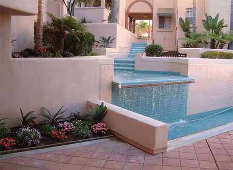 Outdoor courtyard area with a tiered water feature cascading into a pool, surrounded by pink stucco walls, tiled flooring, and various green plants and flowers.