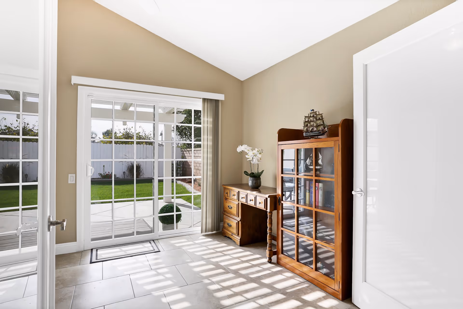 A bright room with beige walls and tiled floor featuring a wooden desk with drawers and a glass-front cabinet displaying books and decorative items. A model ship is placed on top of the cabinet. Large sliding glass doors with white frames open to a green backyard with a white fence and some plants. Sunlight casts shadows on the floor through the glass doors.