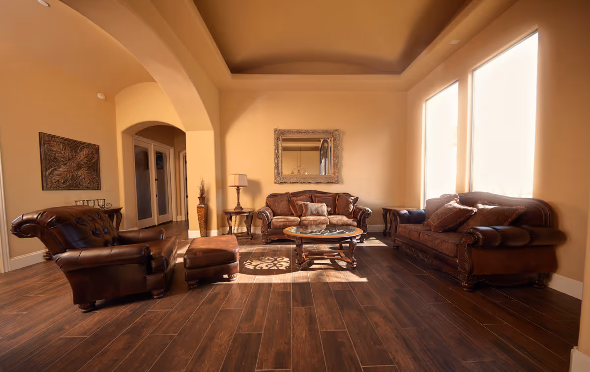 A warm, inviting living room with brown leather sofas and an armchair arranged around a round wooden coffee table. The room features wooden flooring, beige walls, a large decorative mirror above the central sofa, and two tall windows letting in natural light. There is an archway leading to another area and decorative items including a floor vase and a table lamp.